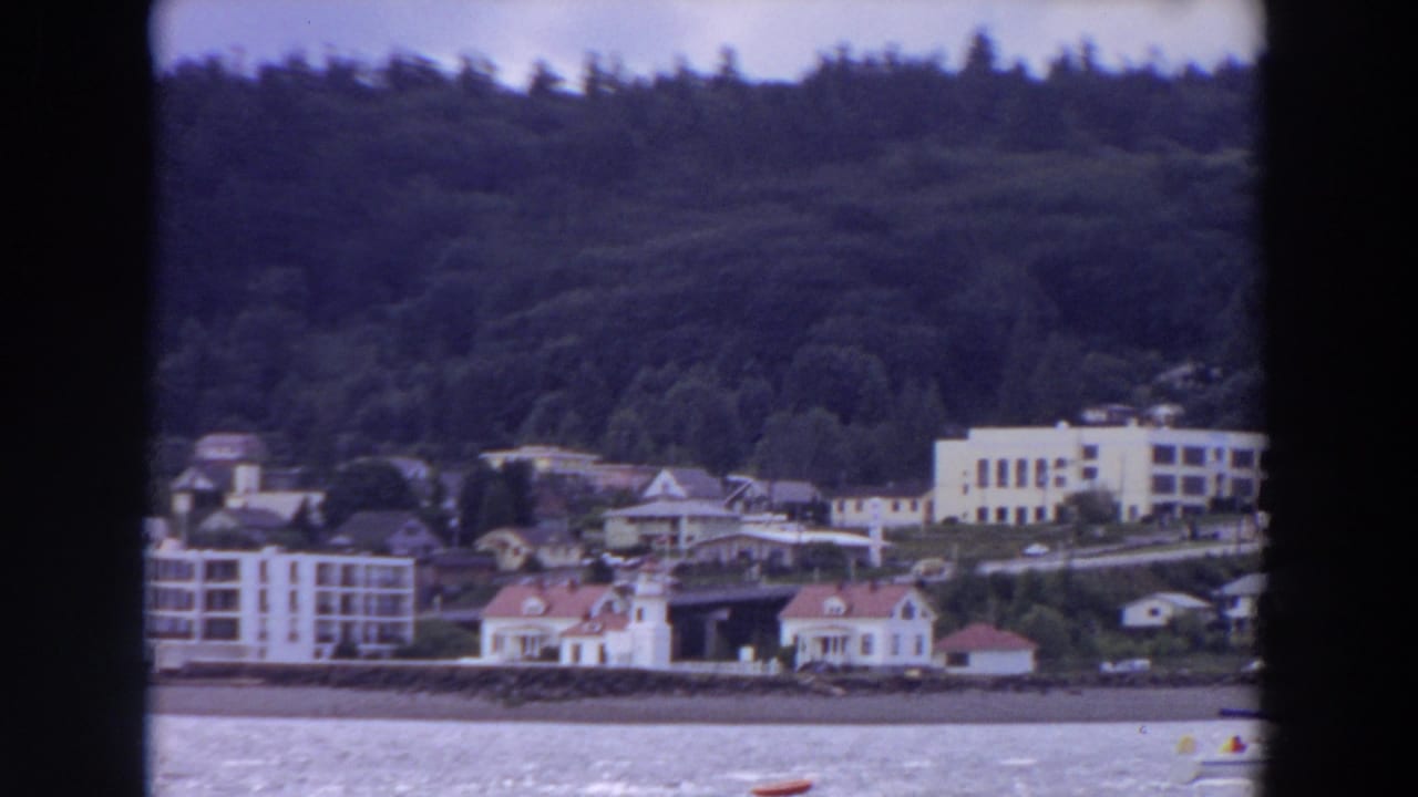 Waterfront town buildings below forested hills, seen across choppy water through dark film frame edges.