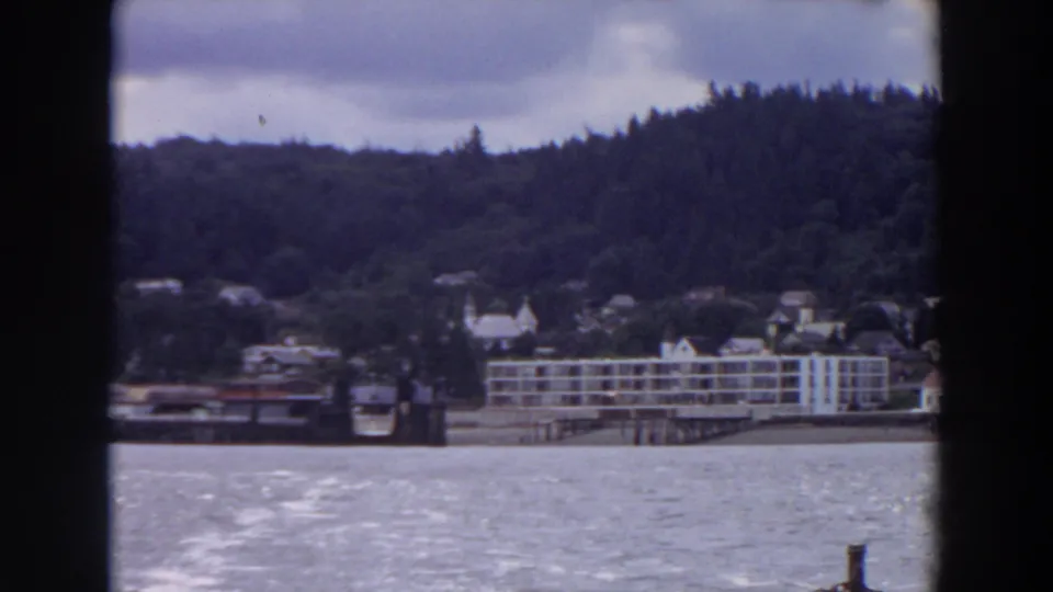 The 8mm view shifts along the waterfront, showing clustered buildings, a pier-like structure, and wooded hills beyond the lake.