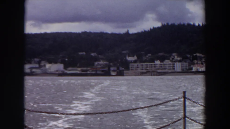 A boat-side 1964 Washington view looks over lake water toward a distant shoreline town, with chain railing in the foreground.