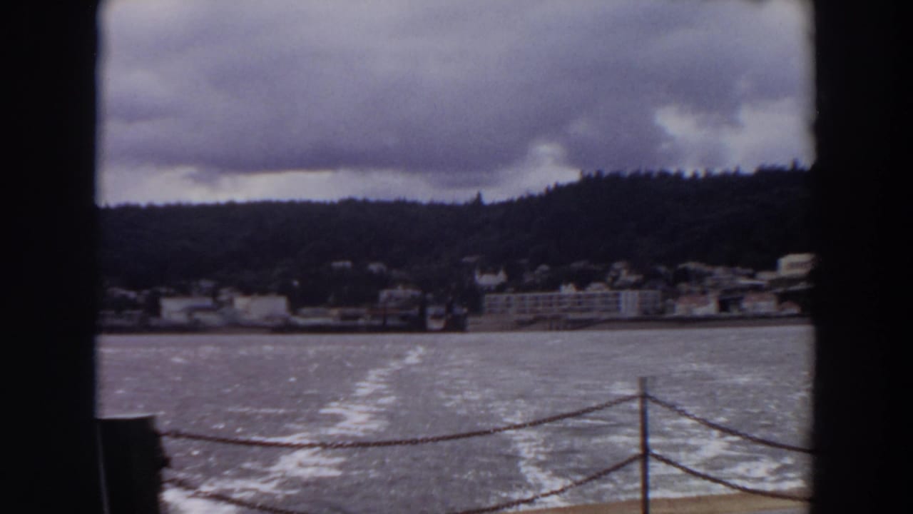 1964 Washington 8mm frame showing a boat wake, deck railing, shoreline buildings, wooded hill, and cloudy sky.