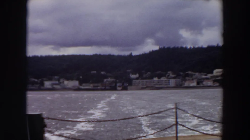 1964 Washington 8mm frame showing a boat wake, deck railing, shoreline buildings, wooded hill, and cloudy sky.