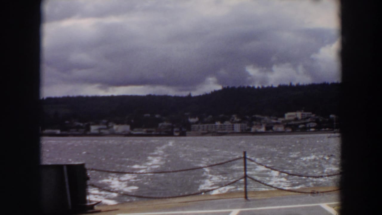 Waterfront recedes behind a moving boat, framed by dark film edges and deck railing.