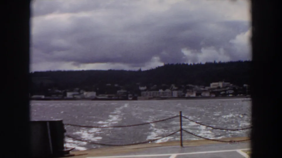 1964 Washington home-movie view from a boat deck with wake, chain railing, shoreline buildings, wooded hill, and clouds.