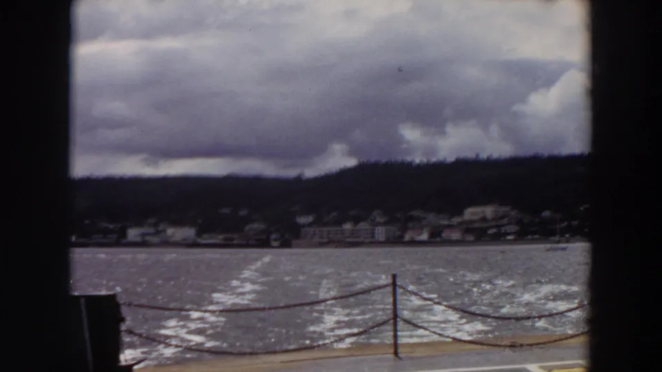 1964 Washington 8mm still with chain railing, deck edge, choppy wake, waterfront buildings, hill, and overcast sky.
