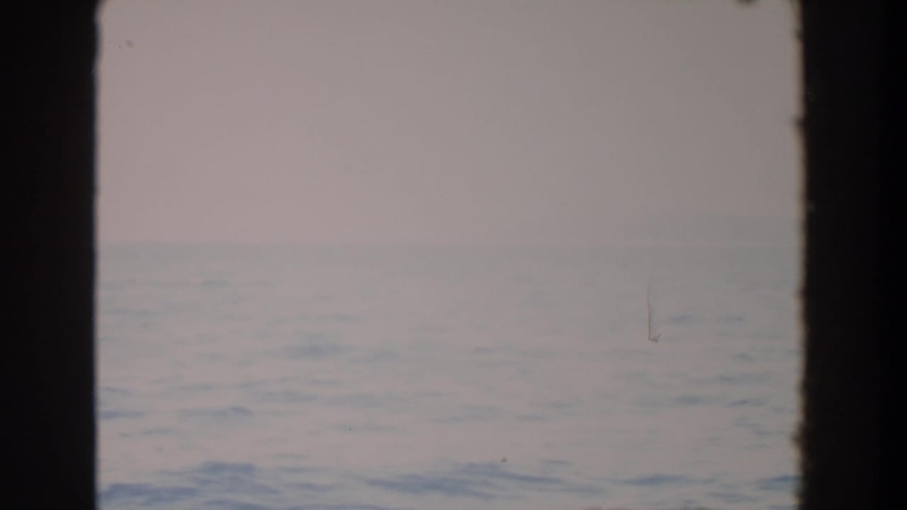 1964 Washington-associated 8mm frame showing a muted ocean horizon through dark window mullions, with soft sky and visible film grain.