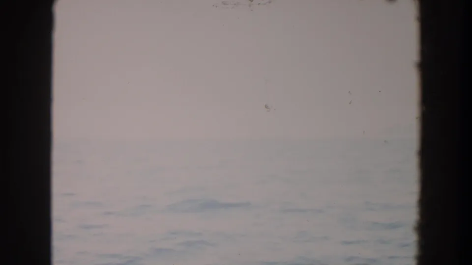 A soft 8mm view of open water under a pale sky, split by a dark vertical window mullion with dust marks visible on the scan.
