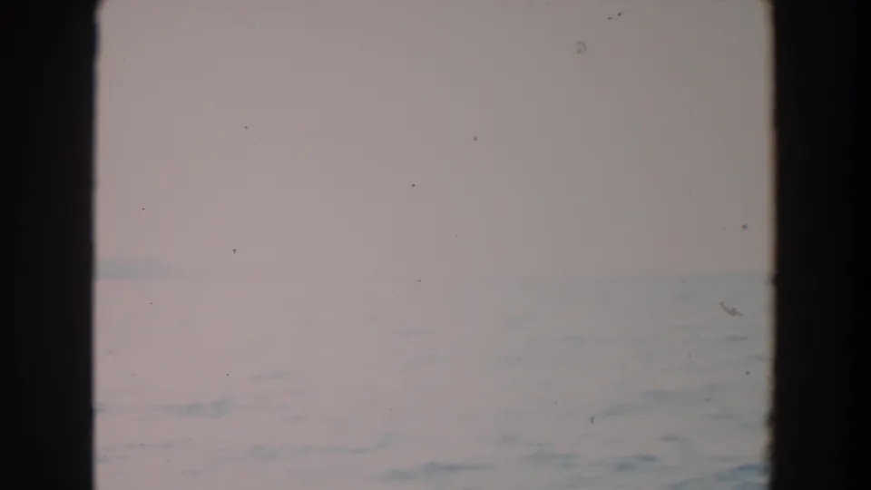 The final 8mm frame holds on a distant ocean horizon through dark window framing, with pale overcast light and minor film specks.