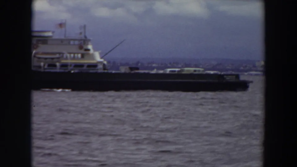 Side view of the vessel traveling across the river, with an American flag and distant waterfront visible through soft 8mm grain.