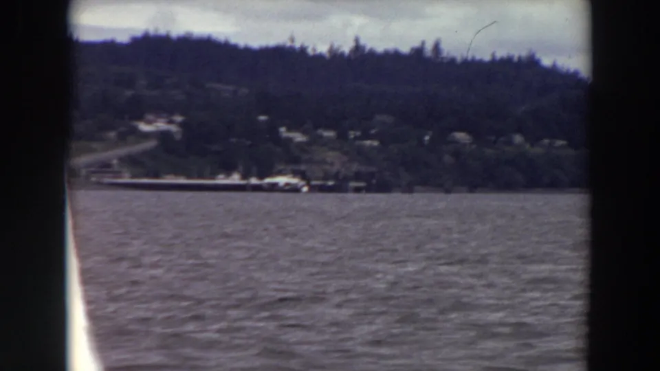 1964 Washington shoreline view with a wooded hill, scattered houses or small buildings, and gray water in the foreground.
