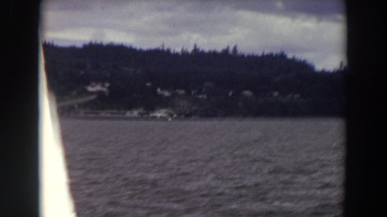 1964 Washington 8mm frame from a moving boat, showing dark water, evergreen shoreline, distant houses, and an overcast sky.