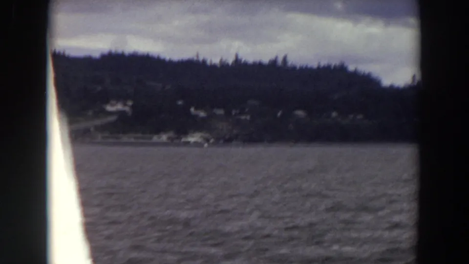 1964 Washington 8mm frame from a moving boat, showing dark water, evergreen shoreline, distant houses, and an overcast sky.