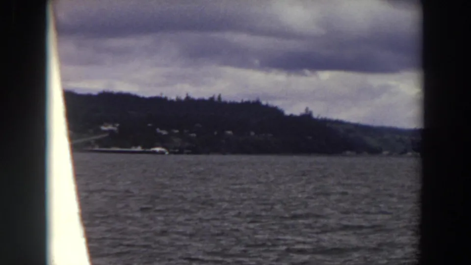 1964 Washington home-movie view from a boat, looking across choppy water to evergreen-covered land and distant shoreline structures.