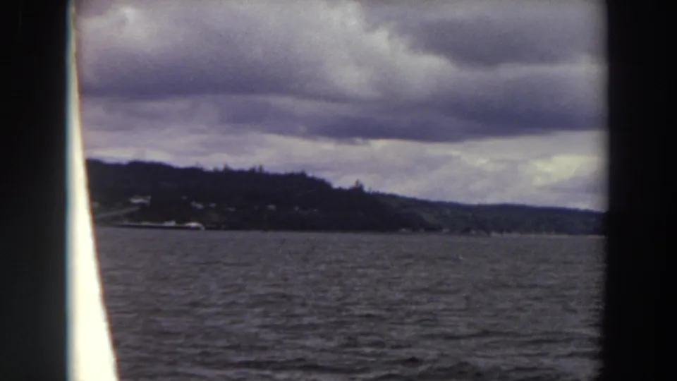 Washington 1964 boat-perspective frame with open water, a wooded shoreline point, distant hills, and heavy overcast clouds.