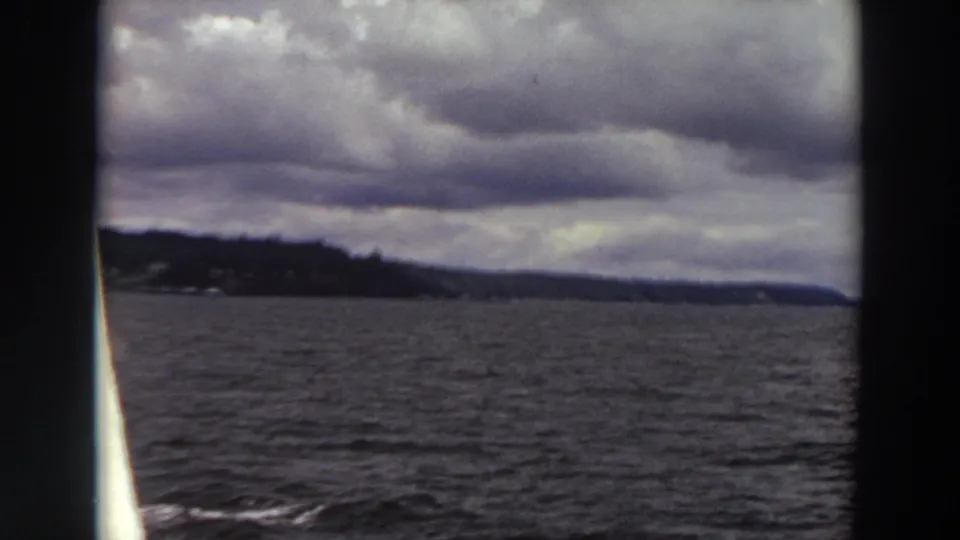 Final 1964 Washington 8mm frame shows rippling water, a low tree-lined shore, distant hills, and dramatic cloud layers from a boat.