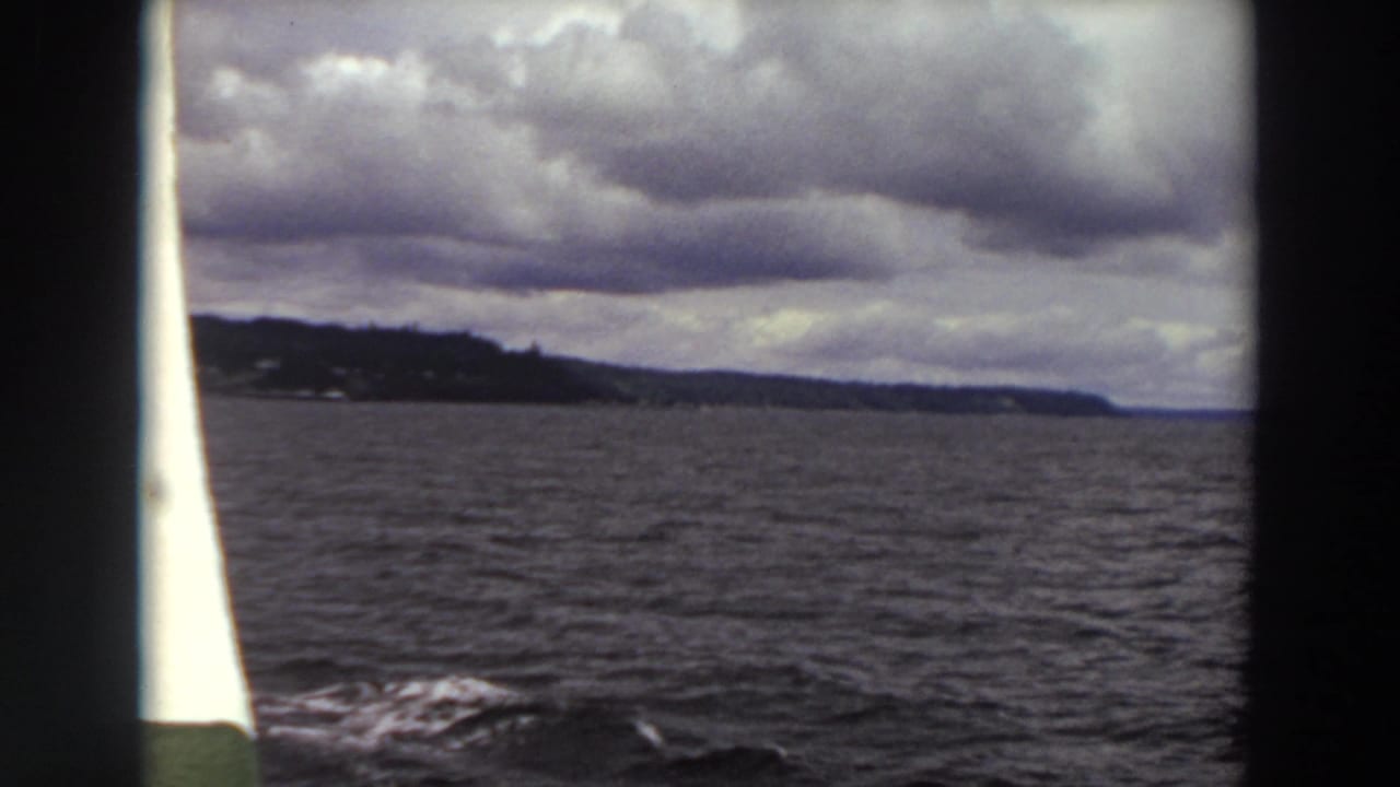 1964 Washington 8mm frame showing choppy water and a distant wooded shoreline seen between dark boat edges under layered clouds.