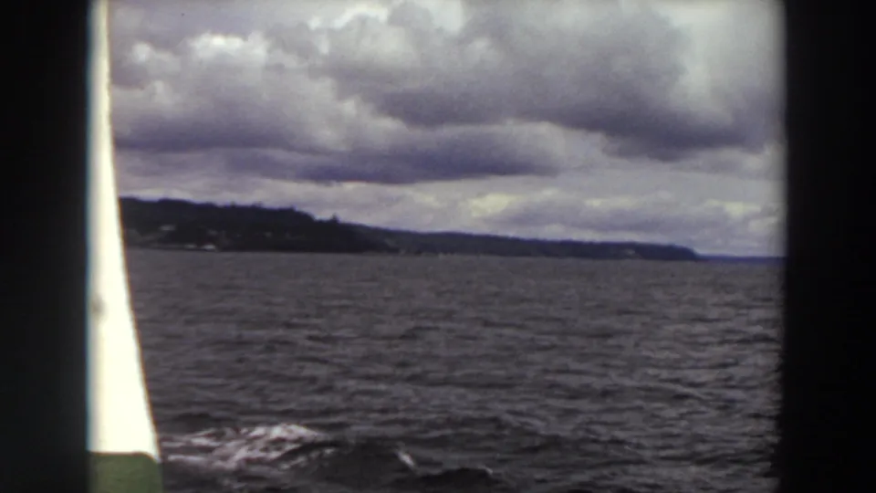1964 Washington 8mm frame showing choppy water and a distant wooded shoreline seen between dark boat edges under layered clouds.