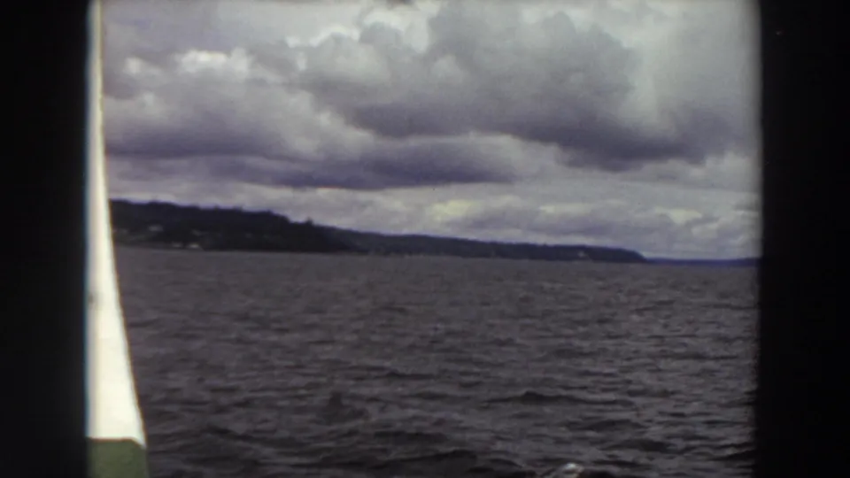 Washington 1964 home-movie frame with rippled water, low tree-covered land, and overcast sky seen from a boat.