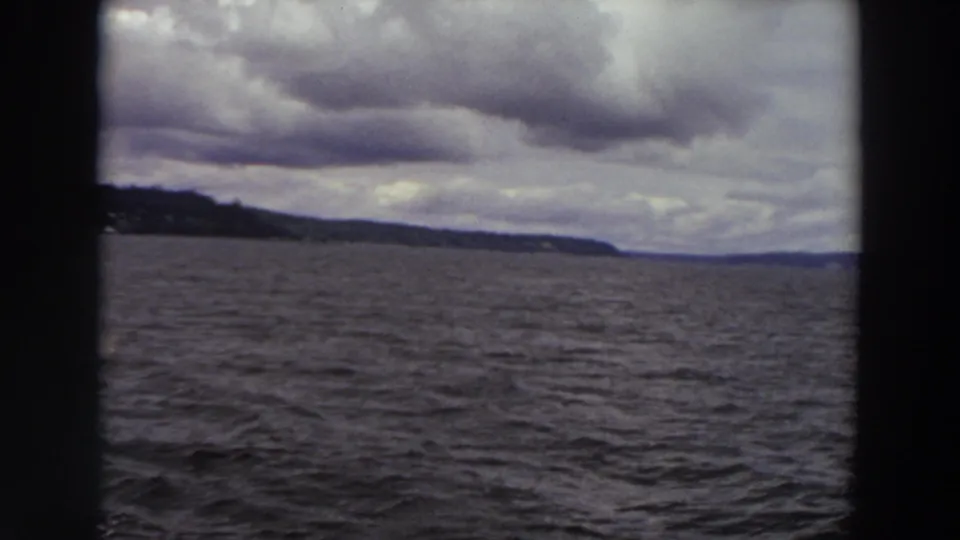 1964 Washington 8mm still of choppy water and a distant low shoreline beneath heavy gray clouds, filmed from a moving boat.