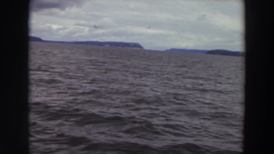 8mm home-movie frame of open water, distant Washington shoreline, and pale clouds from a vessel perspective.