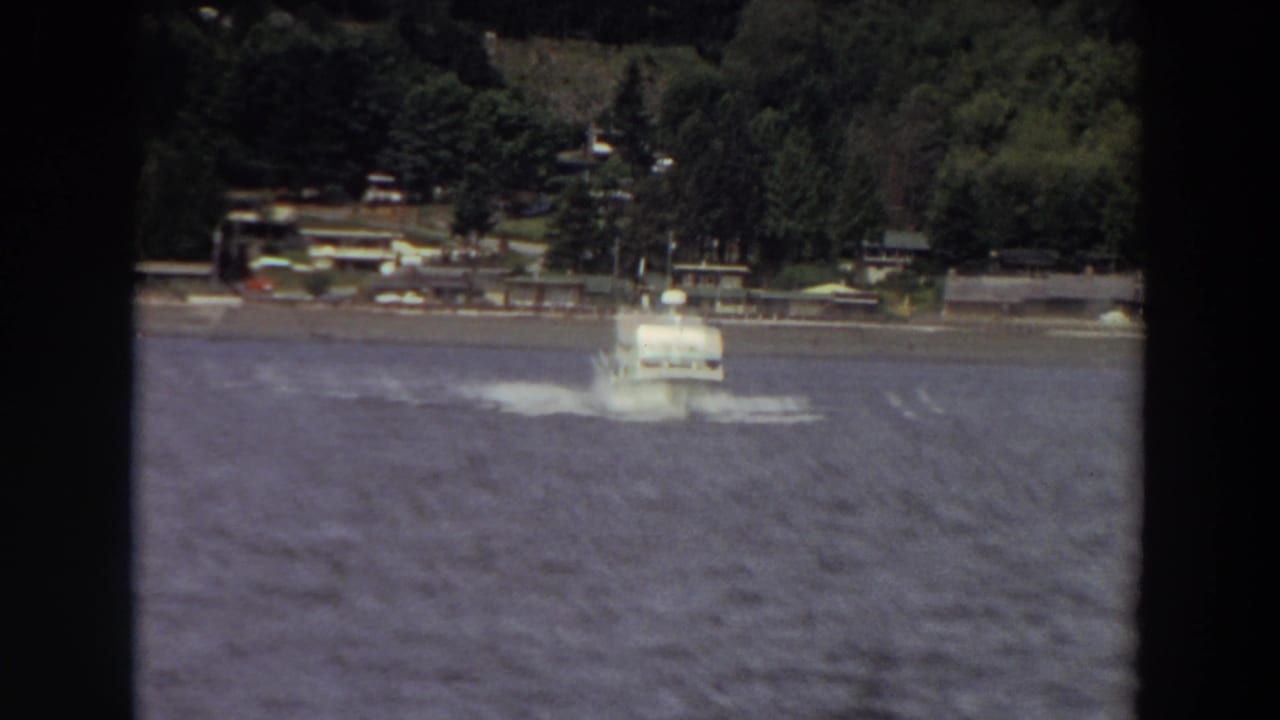 1964 Washington home-movie frame showing a small white motorboat on rough water with trees and shoreline structures behind it.