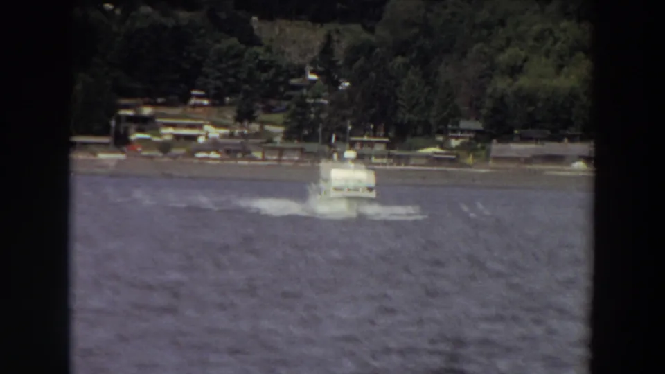 1964 Washington home-movie frame showing a small white motorboat on rough water with trees and shoreline structures behind it.