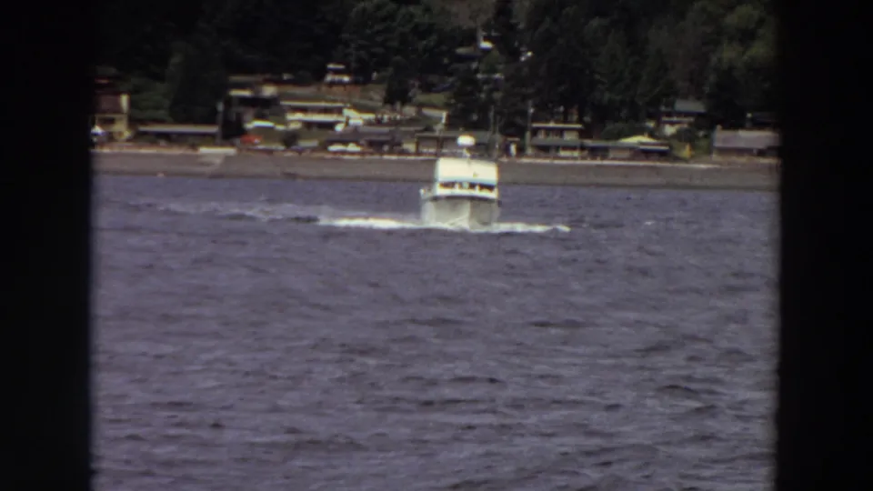 White motorboat on choppy Washington water in a 1964 8mm frame, with waterfront buildings and trees in the background.