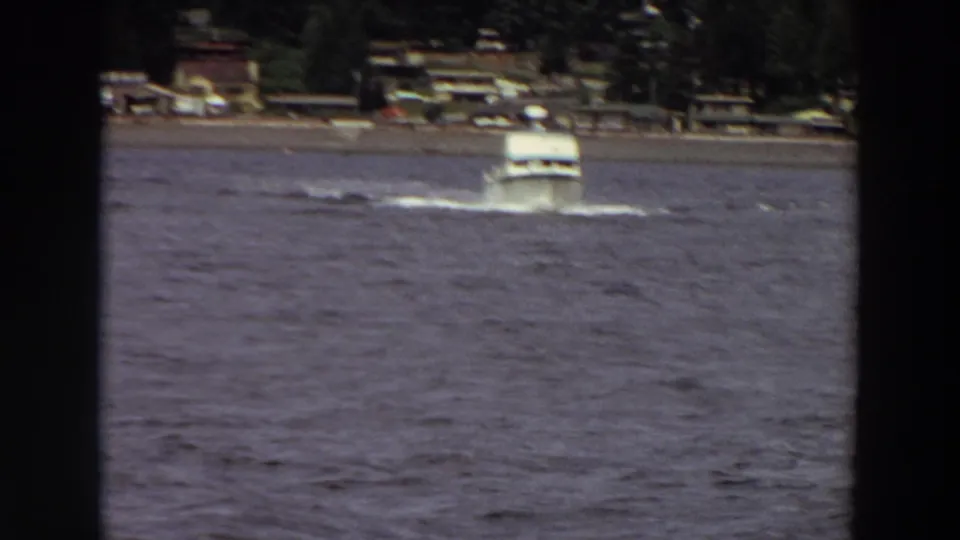 1964 home-movie still of a white motorboat pushing through rough water near a developed, wooded Washington shoreline.