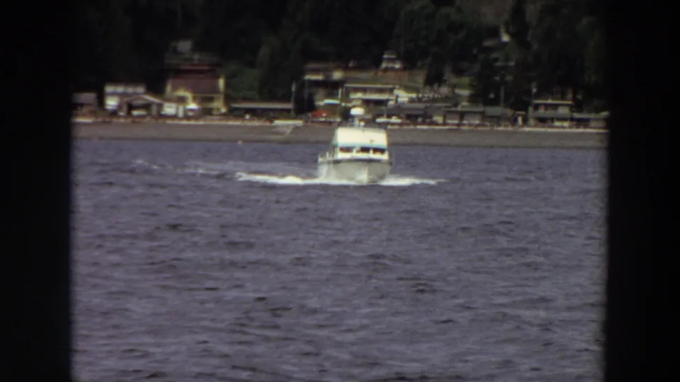 A white motorboat cuts through choppy water near a Washington shoreline, with beach, buildings, and trees visible in this 1964 8mm frame.