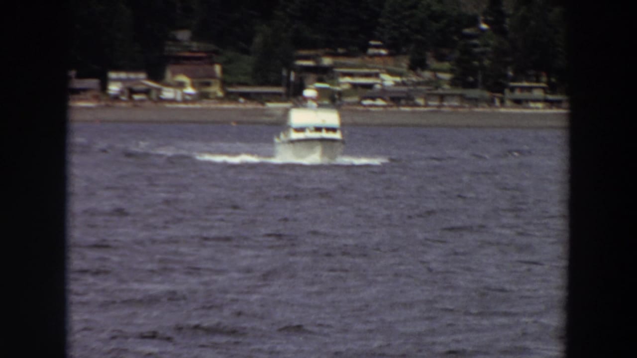 A distant white cabin cruiser approaches across choppy water in archival 1964 home-movie footage from Washington, with shoreline houses and trees in the background.