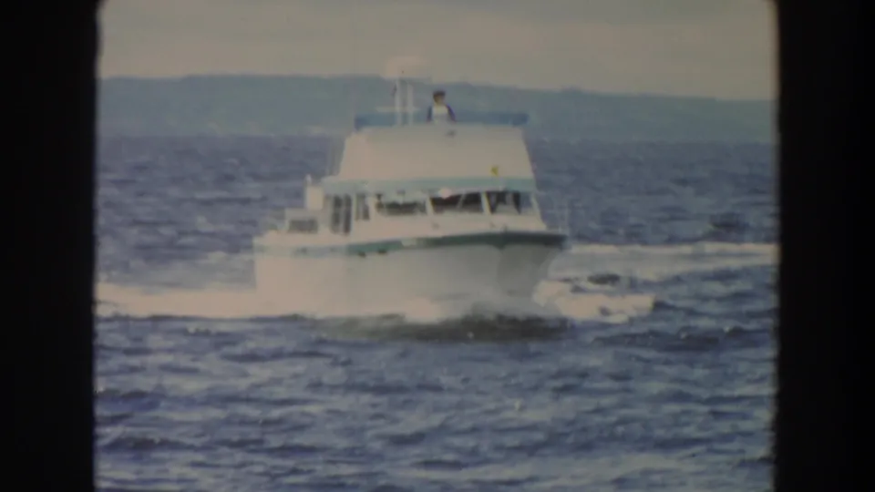 The white cabin cruiser continues across the frame on rough-looking water, with an upper-deck figure and a hazy shoreline beyond.
