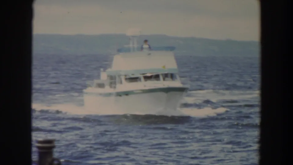 1964 Washington home-movie frame showing a white motor yacht crossing choppy water with distant shoreline behind it.