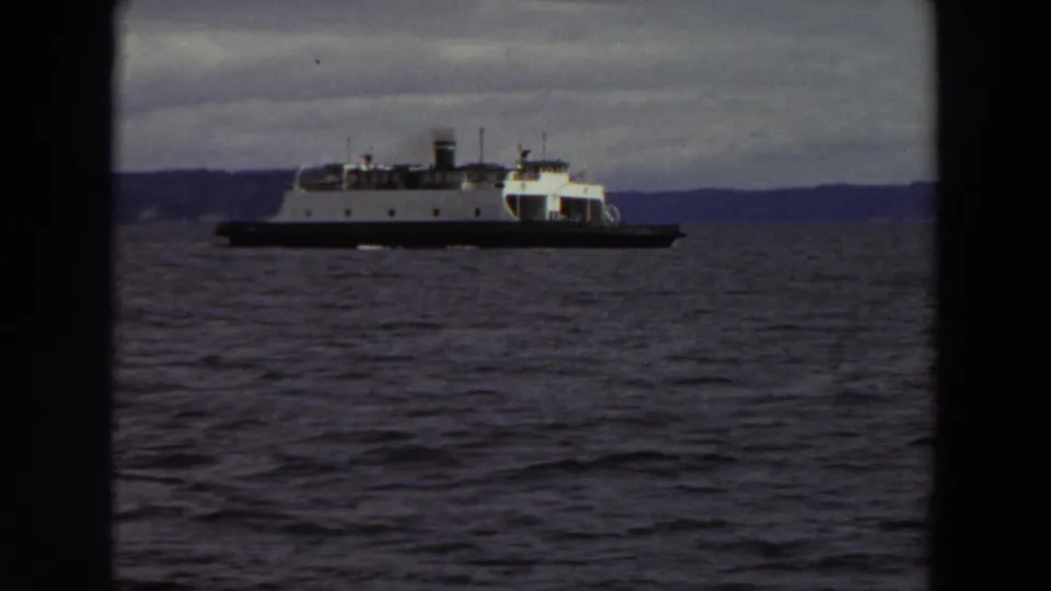A larger industrial ferry or workboat moves across Washington water under a cloudy sky, with low shoreline in the distance.