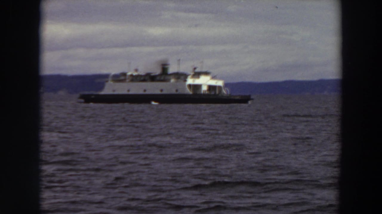 1964 Washington 8mm frame showing a ferry crossing dark water with a low shoreline in the distance under cloud cover.