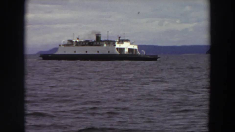 8mm frame from 1964 Washington showing a ferry crossing calm water against a low, clouded shoreline.