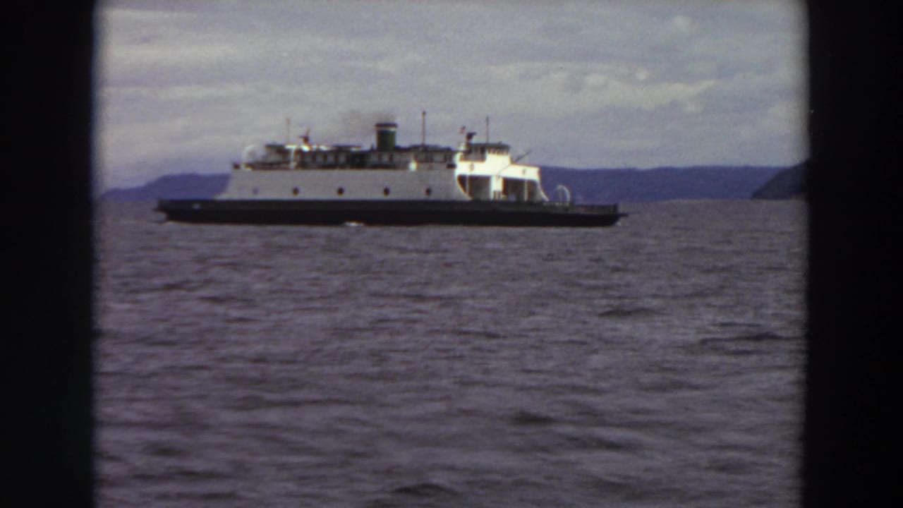 1964 Washington home-movie frame showing a white ferry with a dark hull moving across a waterway, with distant hills beyond.