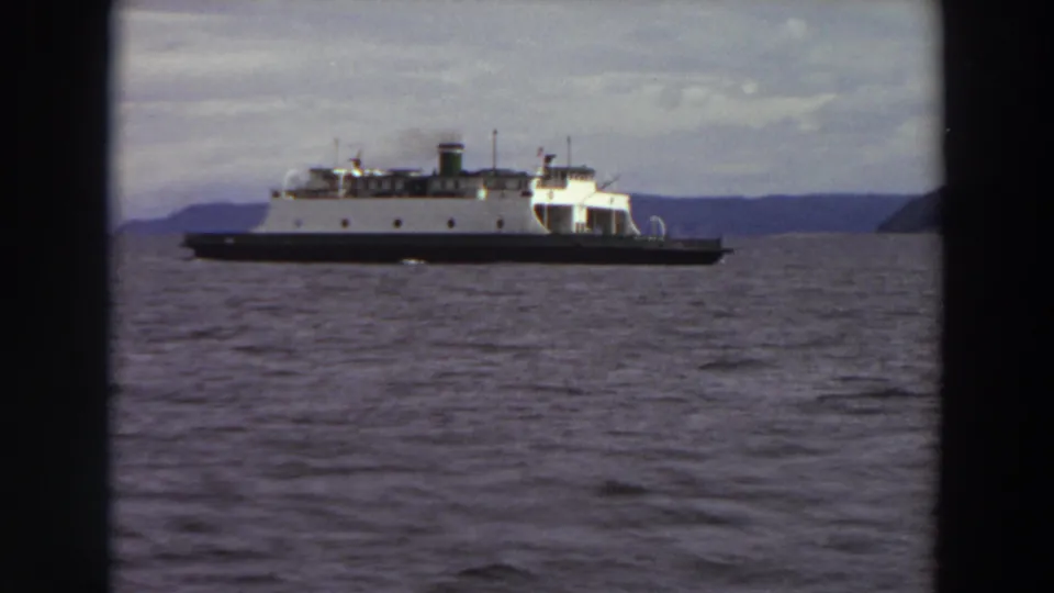 1964 Washington home-movie frame showing a white ferry with a dark hull moving across a waterway, with distant hills beyond.