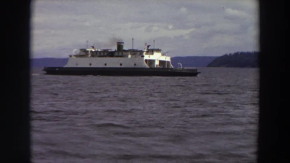 White ferry seen through dark framing as it moves across a Washington waterway beneath a cloudy 1964 sky.