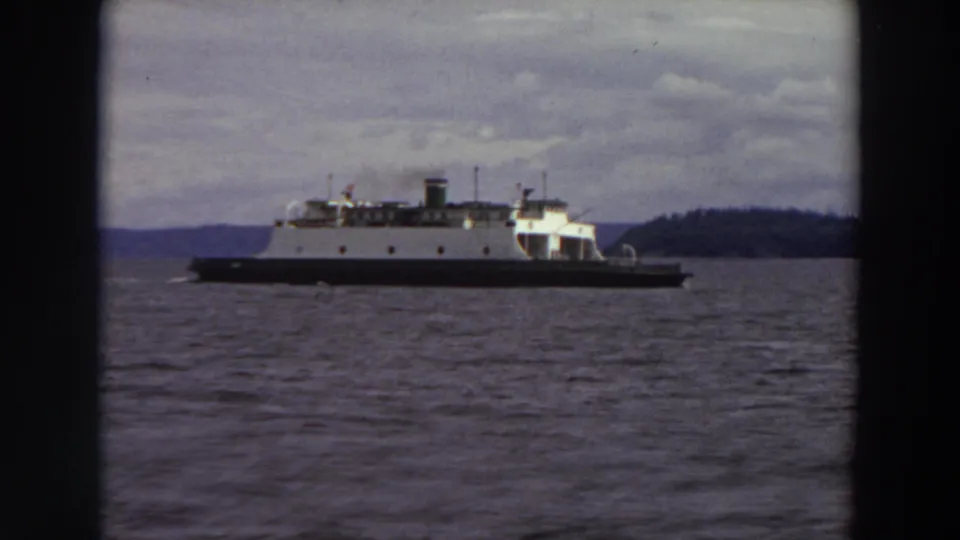 The 1964 8mm frame shows a ferry with smoke from its funnel, dark hull, choppy water, and distant Washington shoreline.