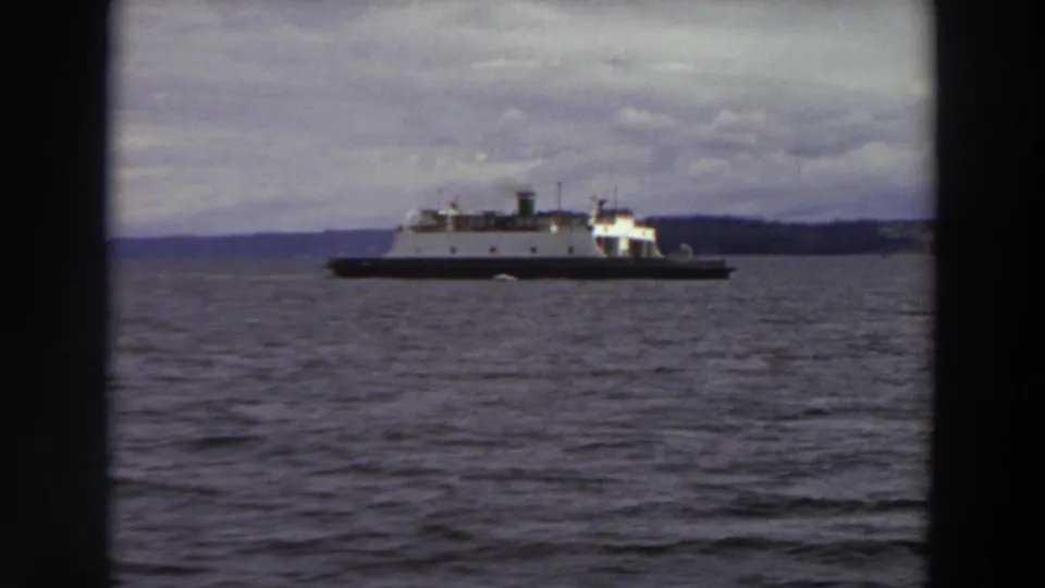 A more distant view of the white ferry crossing a Washington waterway, framed by rippled water, low mountains, and clouds.
