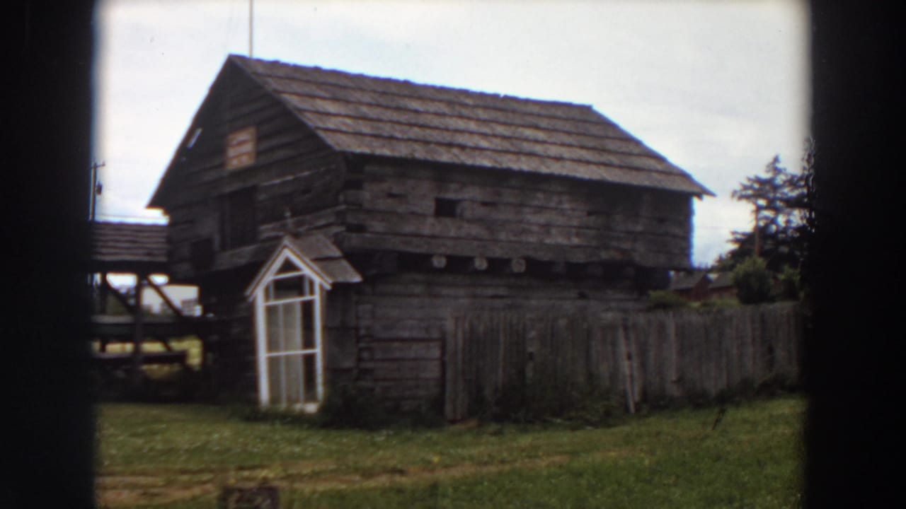1964 Washington home-movie frame showing a rustic wooden cabin-like building with a white door, fence, grass, and overcast sky.