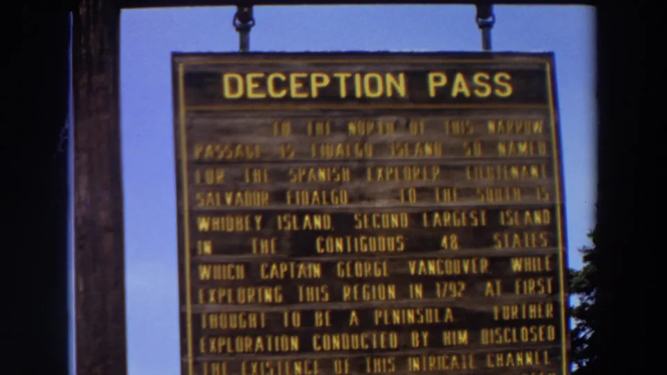 1964 Washington 8mm frame with the hanging Deception Pass sign, support post, blue sky, and a small tree edge.