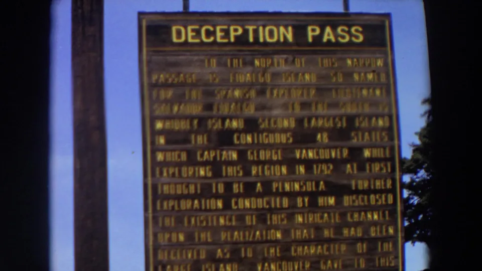 1964 Washington frame centering the wooden Deception Pass informational sign with a pole, blue sky, and trees along the side.