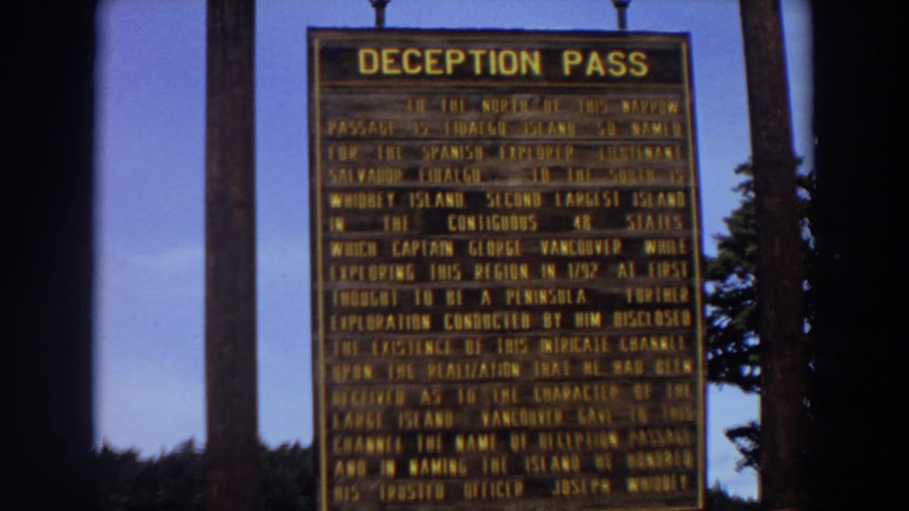 The camera holds on the Deception Pass sign in Washington, with blue sky and trees visible around the marker.