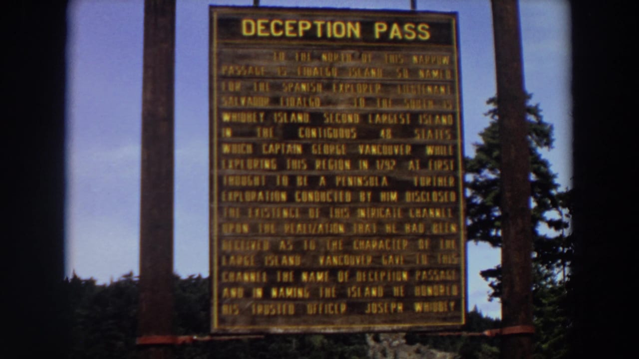 1964 Washington 8mm frame showing the Deception Pass marker centered below blue sky, with wooden supports and trees nearby.