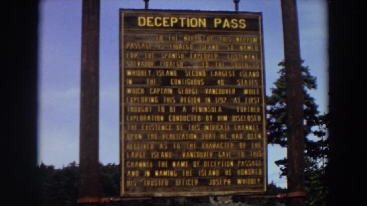 1964 Washington 8mm frame of the Deception Pass sign, with dark film-edge framing, wooden posts, trees, and clear sky.