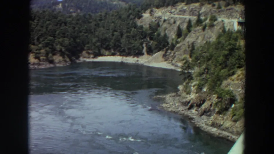 1964 Washington 8mm frame showing a dark river bend bordered by evergreen hills, rocky banks, and a small pale shoreline.