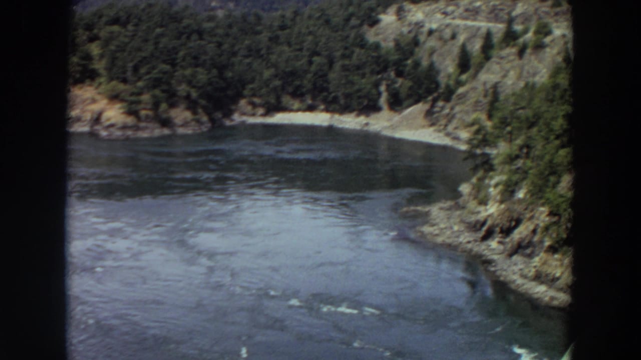 1964 Washington 8mm frame looking down toward rippling water, evergreen-covered banks, rocky slopes, and a small beach.