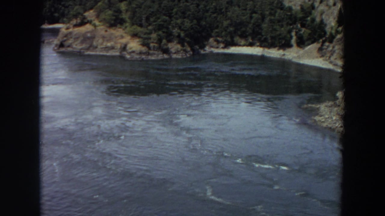 1964 Washington 8mm frame showing a broad river surface beside a wooded point, pale shoreline, and rocky bank.