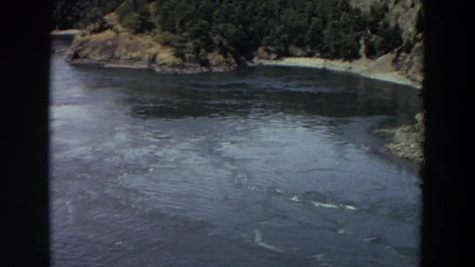 1964 Washington 8mm frame showing a broad river surface beside a wooded point, pale shoreline, and rocky bank.