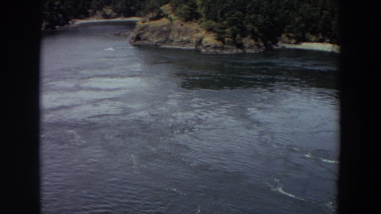 1964 Washington 8mm frame with a wide dark waterway, forested bank, rocky outcrop, and small pale beach in view.